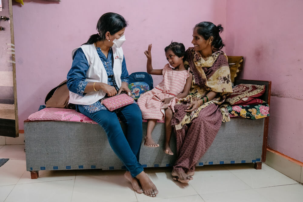 Vaishnavi, de 7 anos, é uma paciente MSF com tuberculose resistente a medicamentos. Na foto, está no colo da mãe, Vishaka, enquanto interage com Prachi, enfermeira MSF.