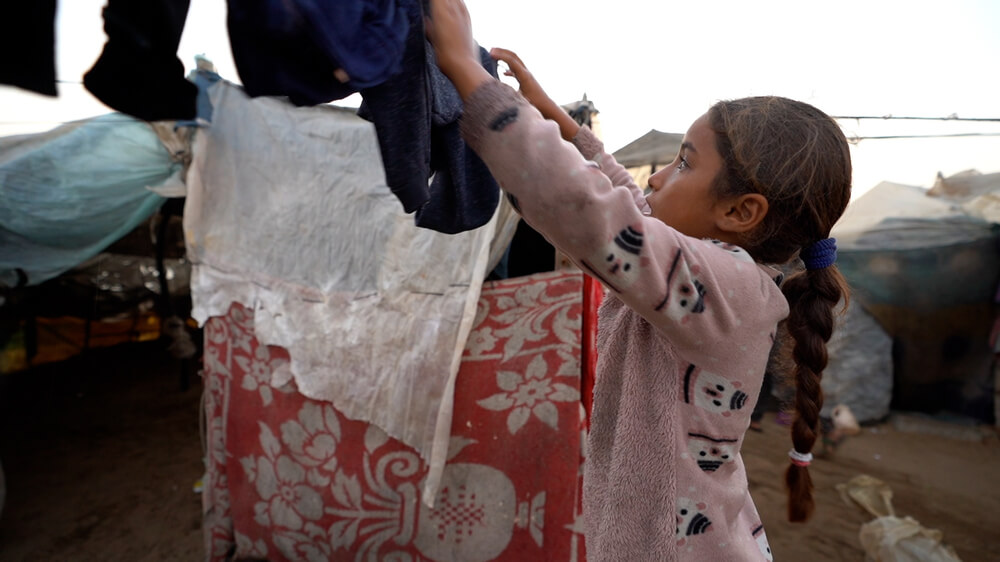 Uma rapariga pendura a roupa da família para secar em frente à tenda onde vivem, em Gaza. Após mais de um ano de guerra e destruição em Gaza, muitas famílias vivem em tendas sobrelotadas ou sob coberturas improvisadas de plástico e tecidos mistos, sem acesso a água limpa, saneamento, produtos de higiene e a outras necessidades básicas.