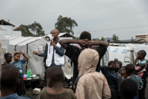 O promotor de saúde da MSF, Aristote Saidi Wanyama, usa um cartaz e um megafone para divulgar informações sobre as estratégias de prevenção da mpox no campo de deslocados de Buhimba, nos arredores de Goma, RDC.