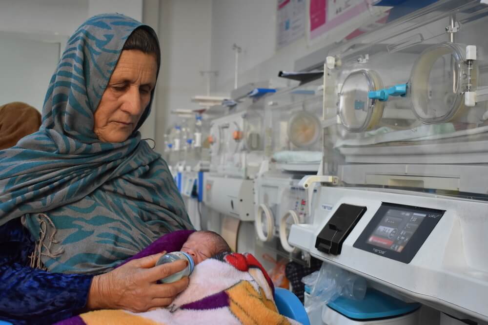Reza Gul, 60 anos, segura o neto nascido prematuramente e o alimenta com leite de uma mamadeira na ala neonatal do Hospital Regional de Mazar-i-Sharif, apoiado pela MSF no Afeganistão. ©️ Logan Turner/MSF