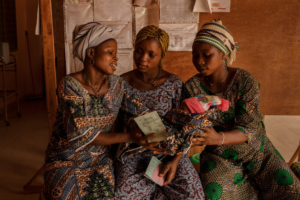 Hortense (à esquerda) entrega o bebé recém-nascido a Melanie (à direita), grávida de seis meses, enquanto aguardam uma consulta no centro de saúde de Klouékanmè. Hortense deu à luz a filha três dias antes, sozinha em casa. Couffo, Benim, janeiro de 2025. Hortense (à esquerda) entrega o bebé recém-nascido a Melanie (à direita), grávida de seis meses, enquanto aguardam uma consulta no centro de saúde de Klouékanmè. Hortense deu à luz a filha três dias antes, sozinha em casa. Couffo, Benim, janeiro de 2025.