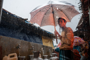 Ershadullah, um refugiado rohingya que fugiu de Myanmar em 2017, abriga-se debaixo de um guarda-chuva durante chuva intensa no campo de refugiados de Kutupalong. Cox’s Bazar, Bangladesh, maio de 2025.